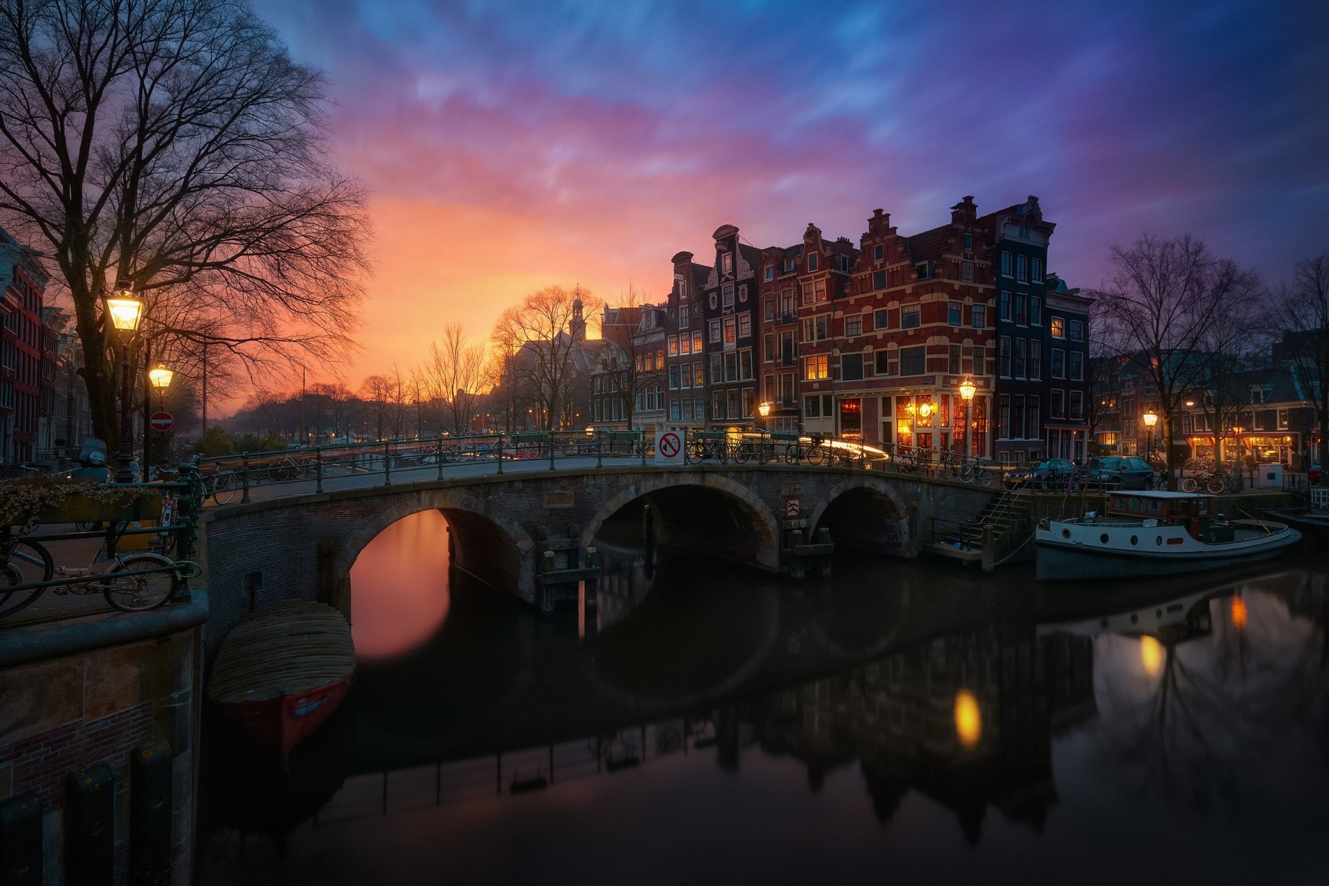 Sunset view of a historic Amsterdam canal with traditional Dutch buildings and a stone bridge, captured in this HD PC desktop wallpaper showcasing man-made beauty in the Netherlands.