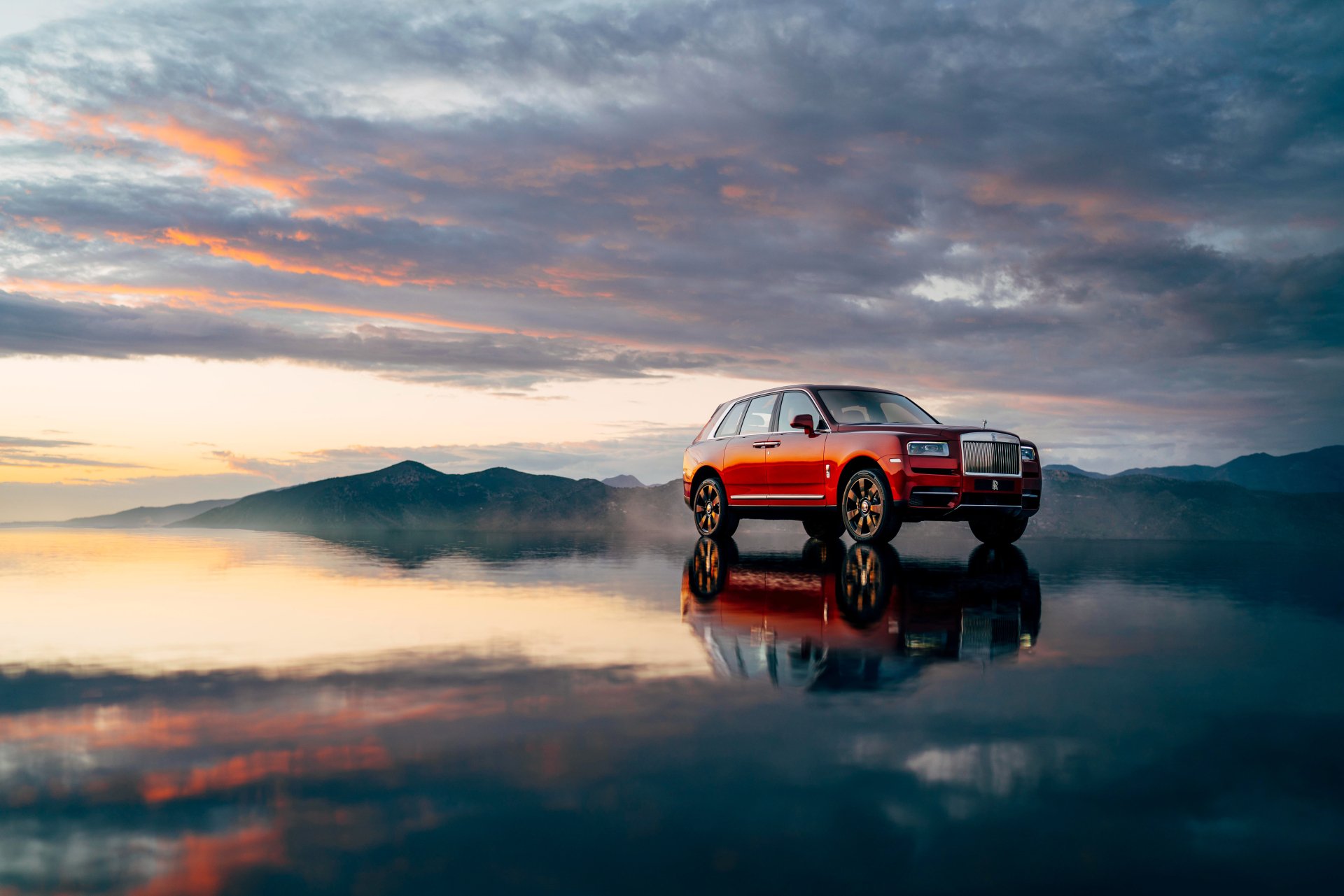 Red Rolls-Royce Cullinan SUV reflected on glassy water under dramatic sunset clouds — 4K Ultra HD PC desktop wallpaper background.