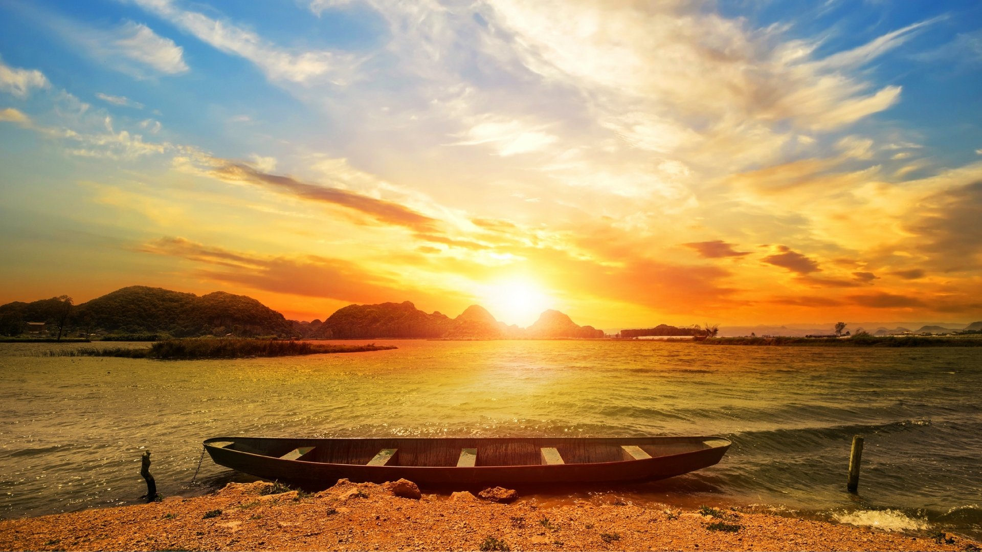 A canoe rests on a beach at sunset, with vibrant clouds and sunshine reflecting over the water under a colorful sky.