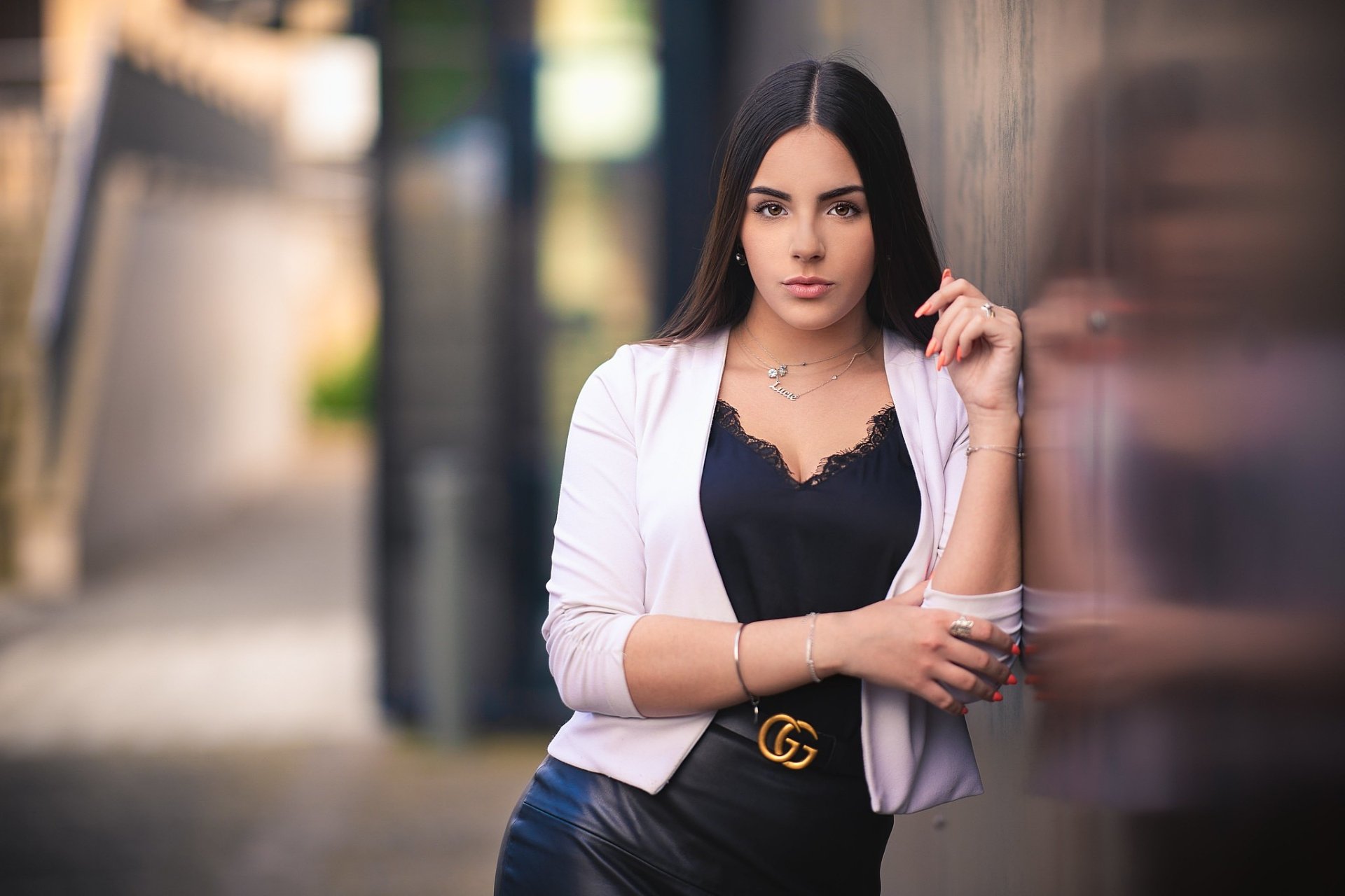 HD desktop wallpaper of a black-haired model with hazel eyes, wearing a white blazer over a black lace top, leaning against a wall with a shallow depth-of-field urban background.