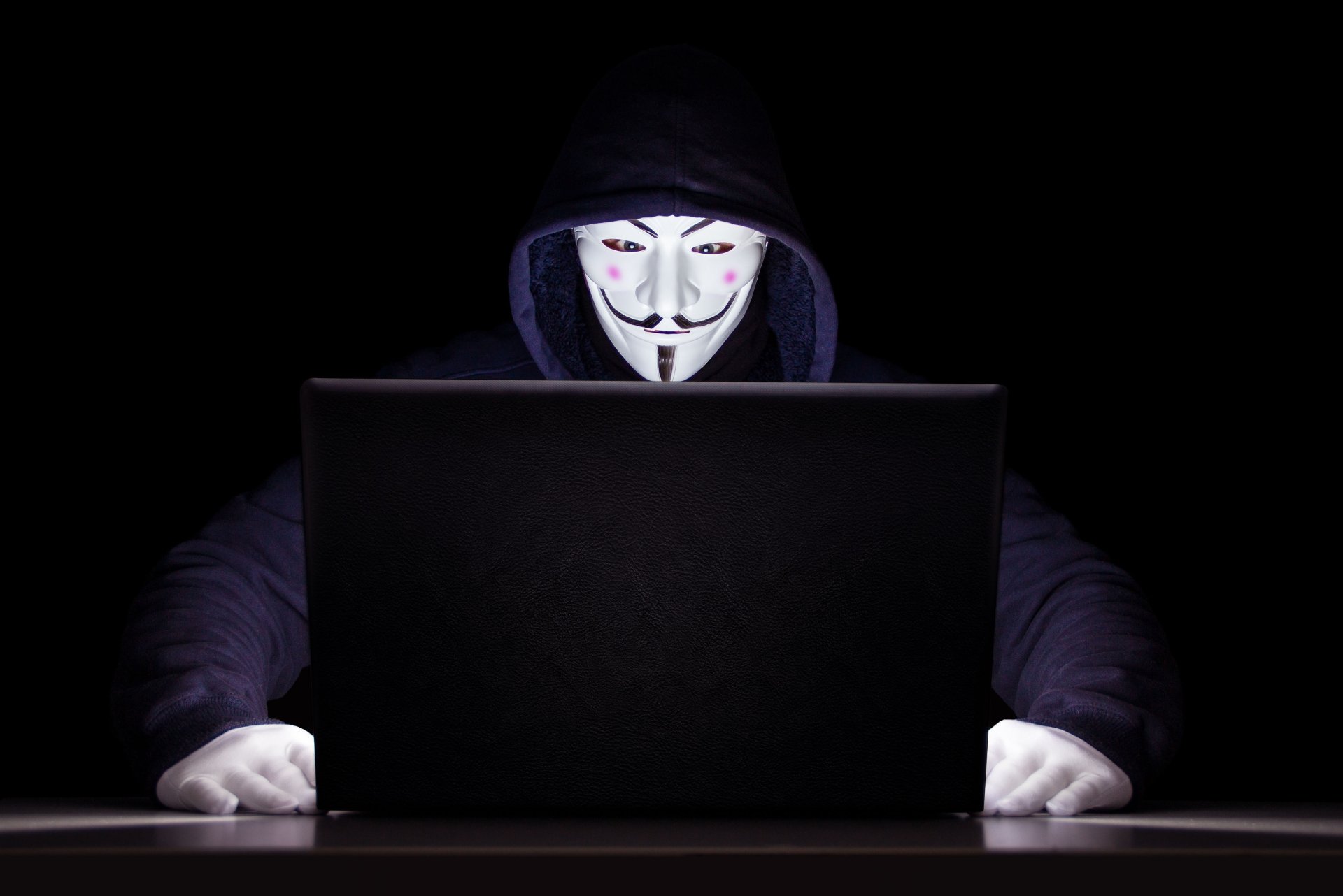Anonymous hacker in a hoodie wearing a Guy Fawkes mask, working on a laptop in a dark setting, captured in 4K Ultra HD for a technology-themed desktop wallpaper.