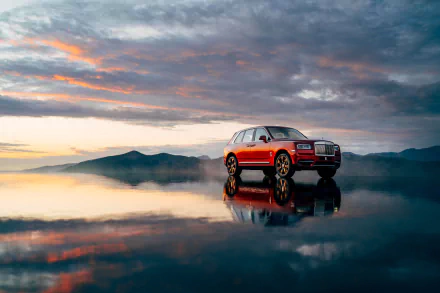 Red Rolls-Royce Cullinan SUV reflected on glassy water under dramatic sunset clouds — 4K Ultra HD PC desktop wallpaper background.