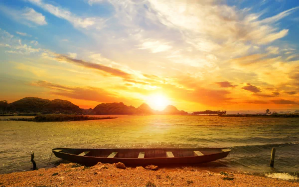 A canoe rests on a beach at sunset, with vibrant clouds and sunshine reflecting over the water under a colorful sky.