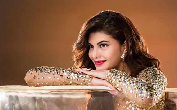 A smiling Jacqueline Fernandez, Sri Lankan actress and model with brunette hair and brown eyes, rests on a glittery surface in this HD desktop wallpaper. She wears red lipstick and a sparkling outfit.