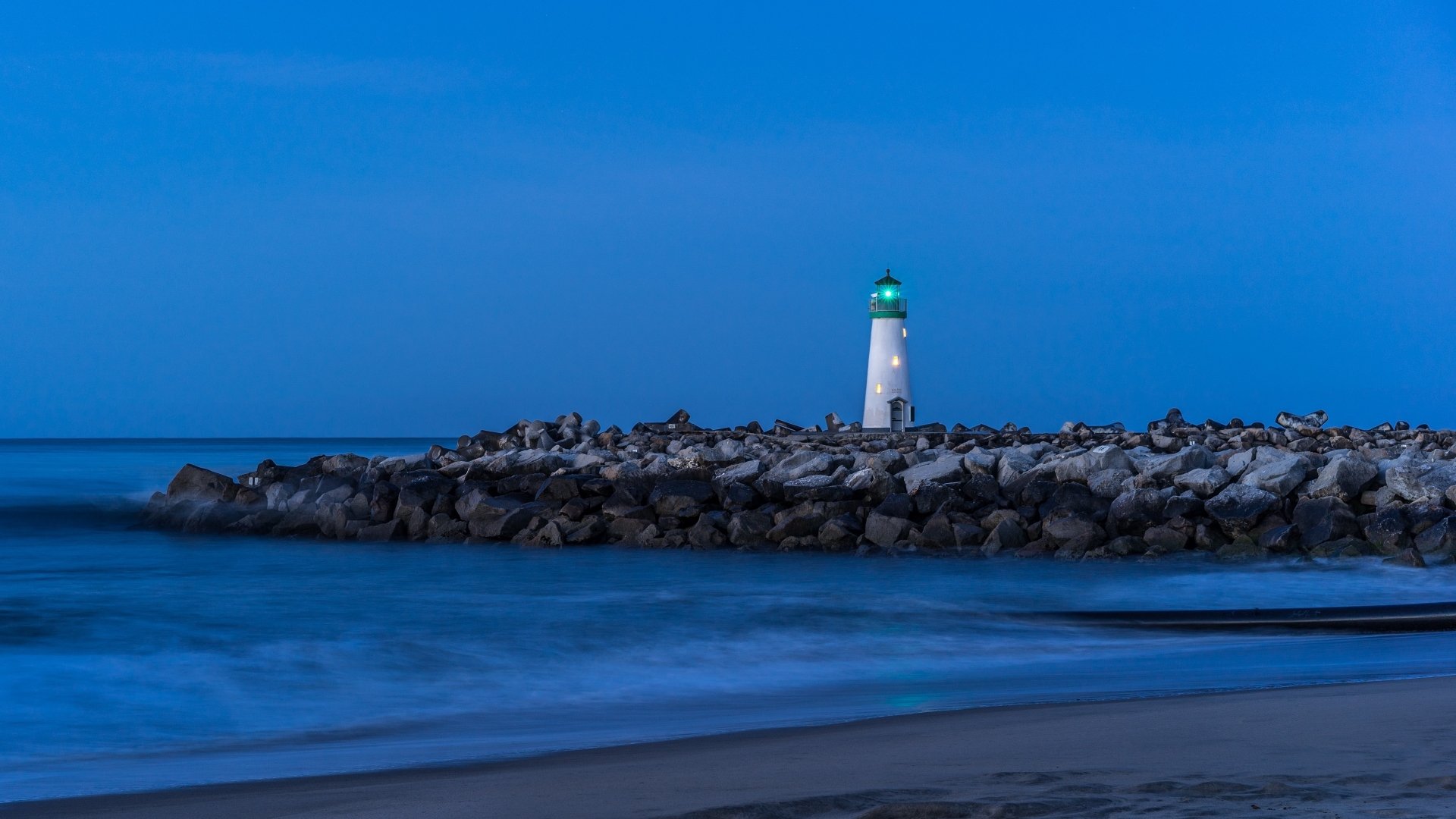 A 4K Ultra HD desktop wallpaper of a white lighthouse glowing at dusk, set against a deep blue sky and calm ocean, with rocky shoreline in the foreground.