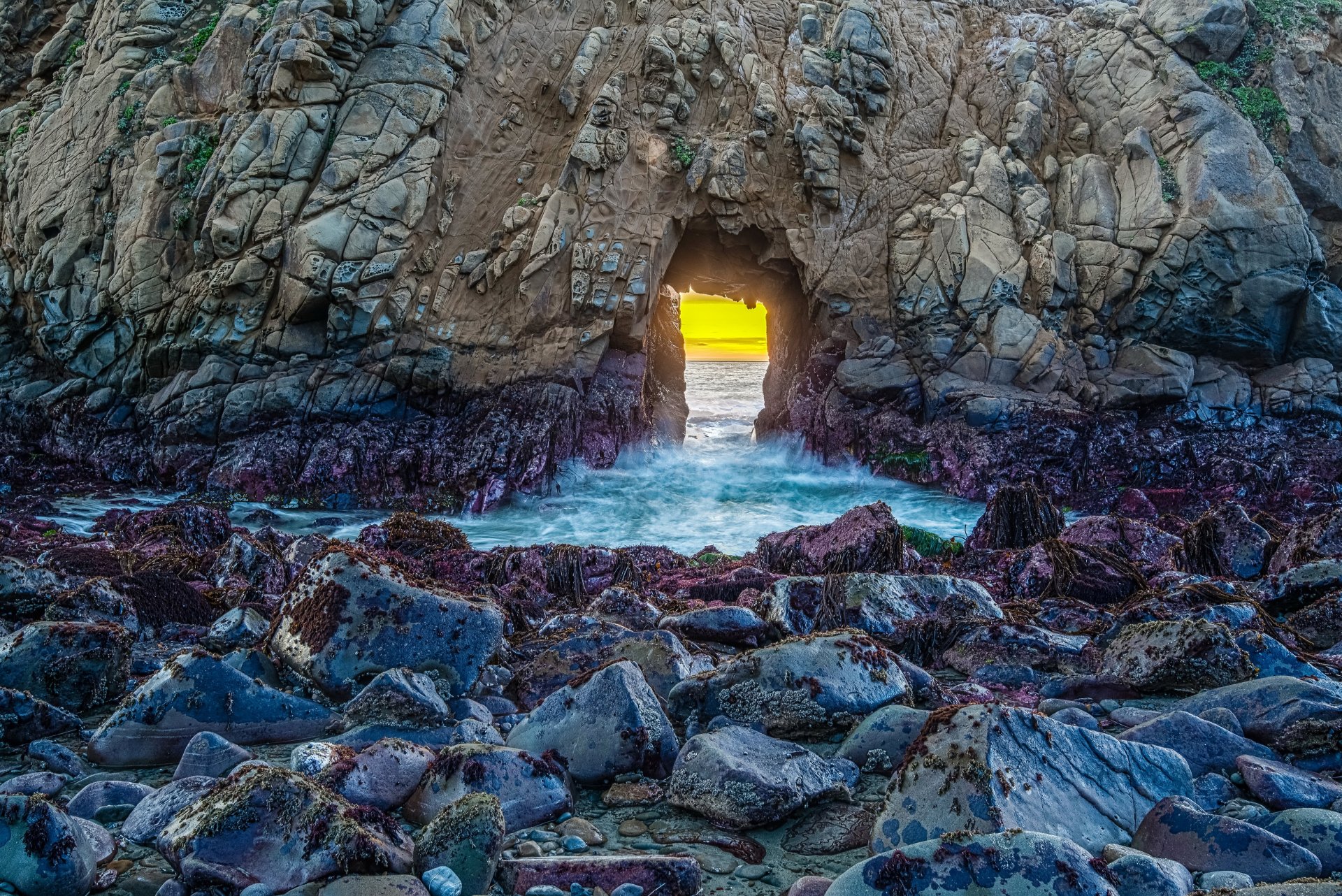 4K Ultra HD desktop wallpaper showing a natural rock arch over rugged coastline with waves crashing through, glowing with golden sunlight in the background.