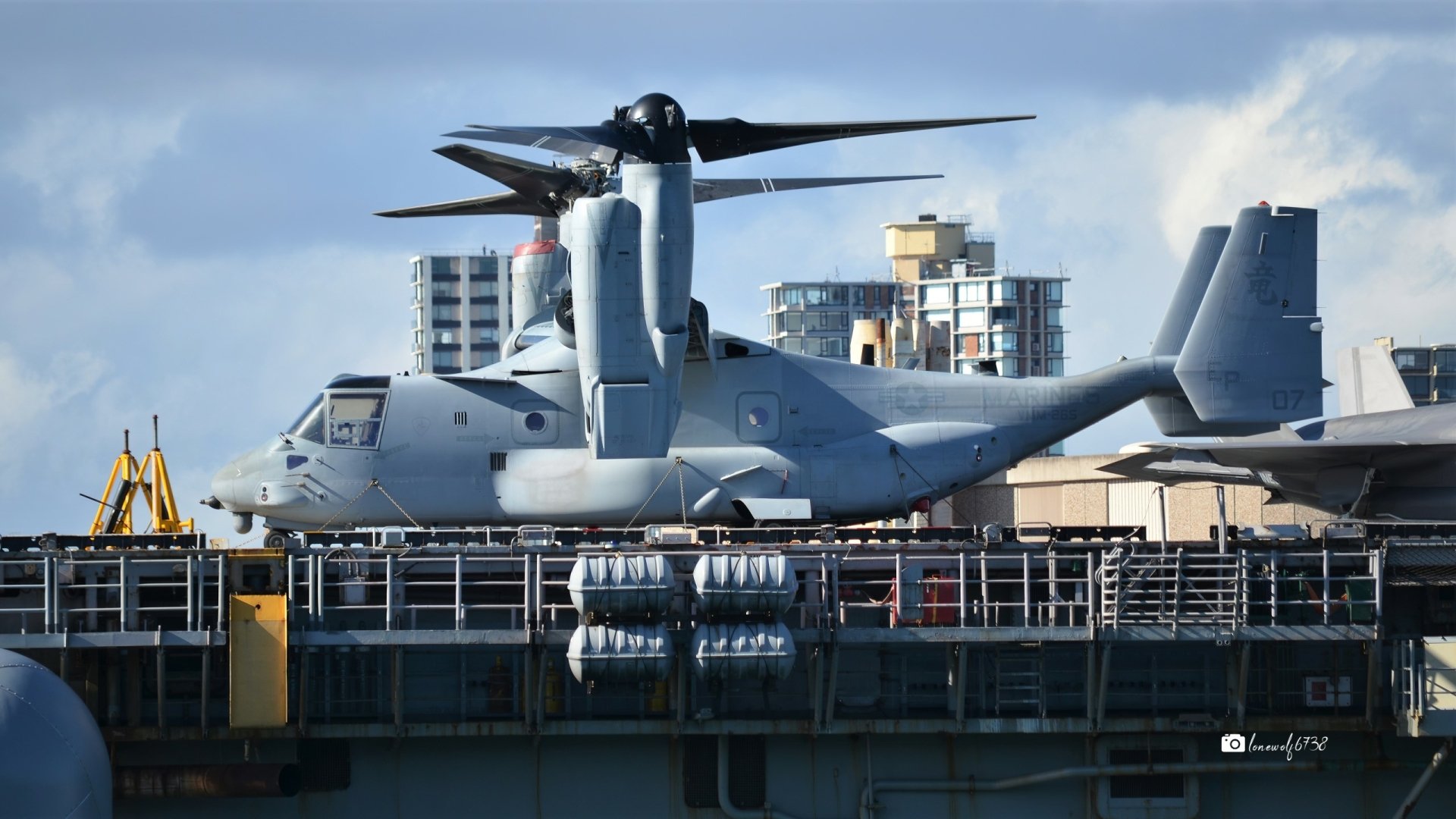 Bell Boeing V-22 Osprey military tiltrotor transport aircraft on a ship deck with city skyline under a cloudy sky — 2K Quad HD PC desktop wallpaper background