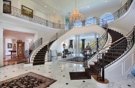A luxurious living room with grand, curved stairs and an elegant chandelier. The high-definition wallpaper showcases a sophisticated interior design with intricate railings and marble flooring.