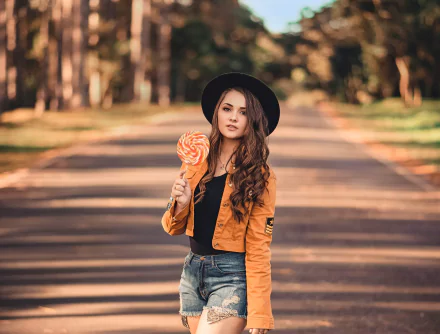 A brunette woman with long hair, wearing a hat, orange jacket, and shorts, holds a large lollipop on a sunlit path with a blurred background, captured in HD.