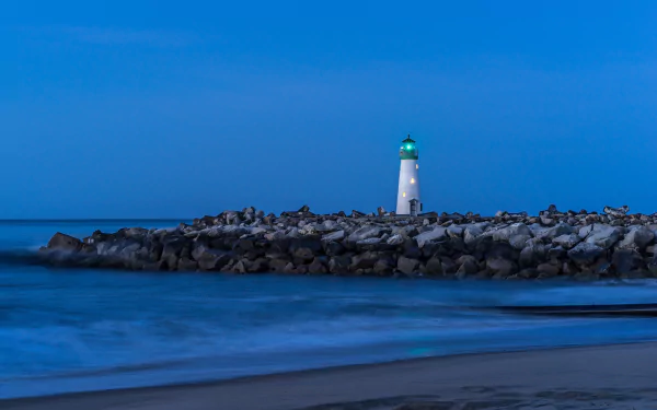 A 4K Ultra HD desktop wallpaper of a white lighthouse glowing at dusk, set against a deep blue sky and calm ocean, with rocky shoreline in the foreground.