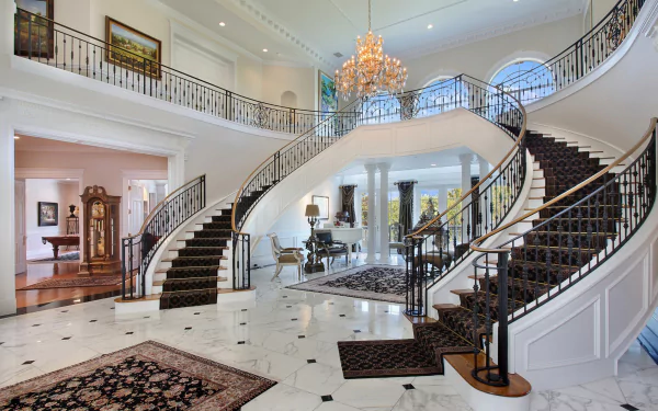 A luxurious living room with grand, curved stairs and an elegant chandelier. The high-definition wallpaper showcases a sophisticated interior design with intricate railings and marble flooring.
