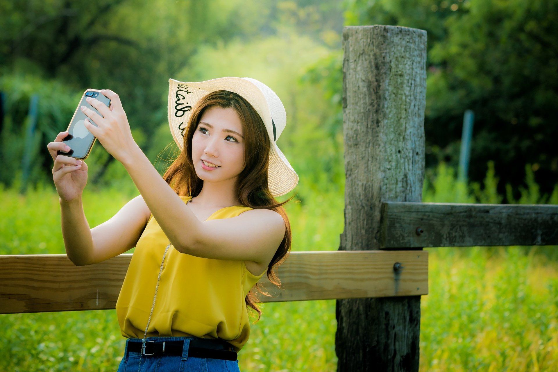 Brunette model with long hair wearing a sun hat, taking a selfie by a wooden fence in a sunlit green field — 4K Ultra HD PC desktop wallpaper.