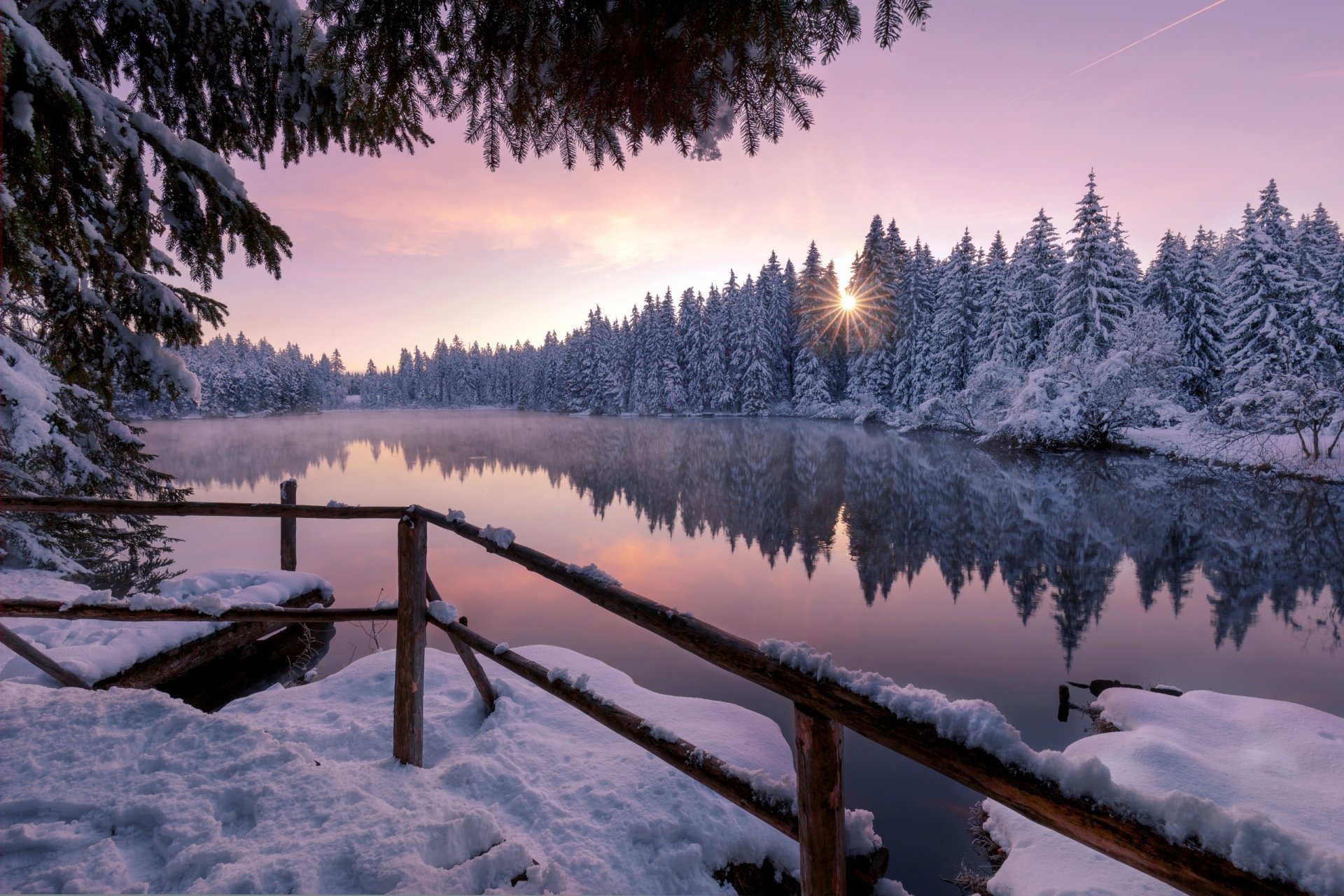 HD desktop wallpaper showing a serene winter landscape with snow-covered trees by a calm river at sunrise, casting soft pink and purple hues across the sky.