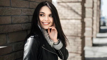 A smiling woman with black hair wearing a leather jacket and gray sweater, posing against a brick wall in a 4K Ultra HD PC desktop wallpaper background.