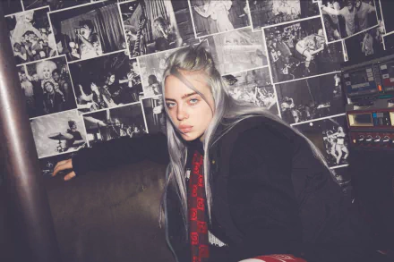 HD desktop wallpaper featuring American singer Billie Eilish with grey hair, set against a wall covered in black-and-white music-themed photos.