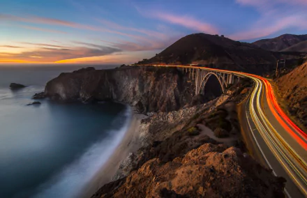 Time-lapse 8K Ultra HD image of California’s Bixby Creek Bridge on a highway at sunset, showcasing vibrant light trails and scenic coastal cliffs.