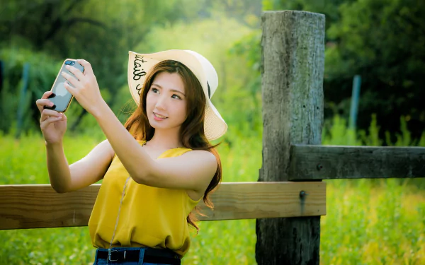 Brunette model with long hair wearing a sun hat, taking a selfie by a wooden fence in a sunlit green field — 4K Ultra HD PC desktop wallpaper.