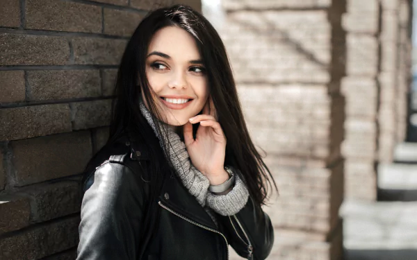 A smiling woman with black hair wearing a leather jacket and gray sweater, posing against a brick wall in a 4K Ultra HD PC desktop wallpaper background.
