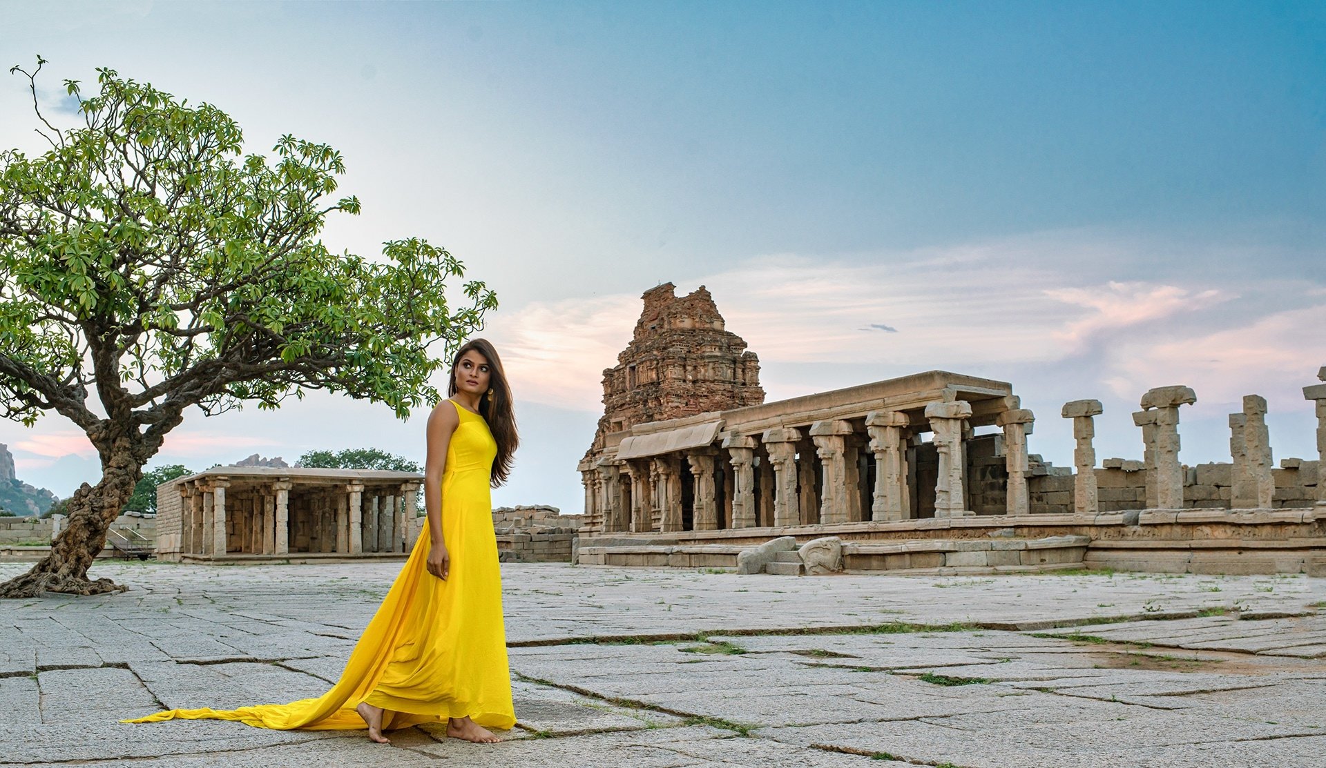 Indian brunette woman in a flowing yellow dress stands near ancient ruins under a clear sky, captured in an HD desktop wallpaper and background.