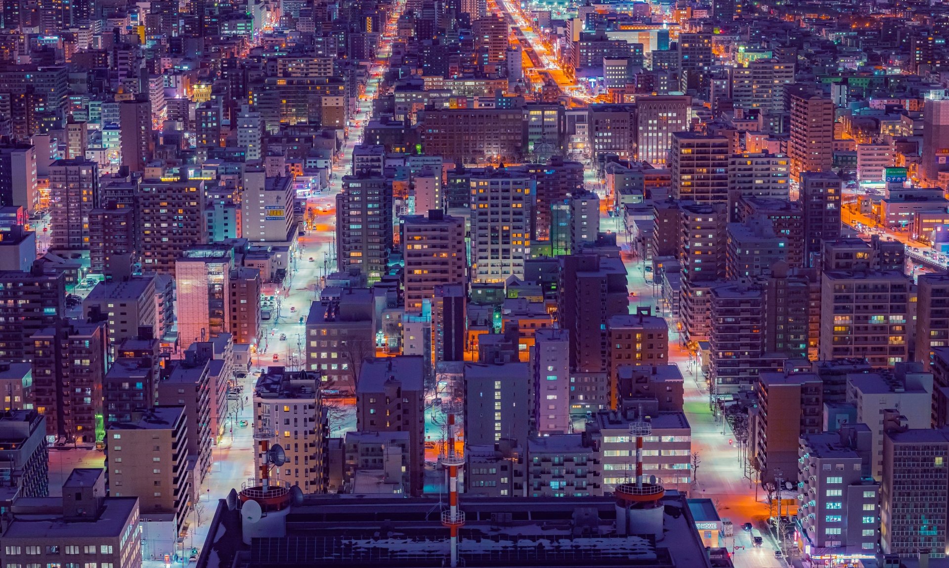 A vibrant 4K Ultra HD man-made cityscape of Sapporo at night, showcasing densely packed illuminated buildings and streets from an elevated perspective.