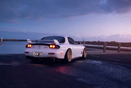 White Mazda RX-7 sports car parked by a waterfront at dusk, rear view with spoiler and gold wheels; 4K Ultra HD PC desktop wallpaper background.