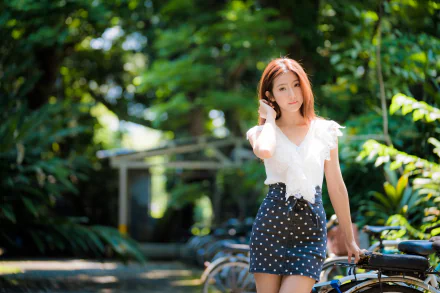 A 4K Ultra HD image of an Asian woman in a polka-dot skirt standing outdoors, captured with a shallow depth of field highlighting her against a lush green background.