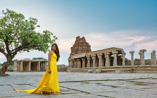 Indian brunette woman in a flowing yellow dress stands near ancient ruins under a clear sky, captured in an HD desktop wallpaper and background.
