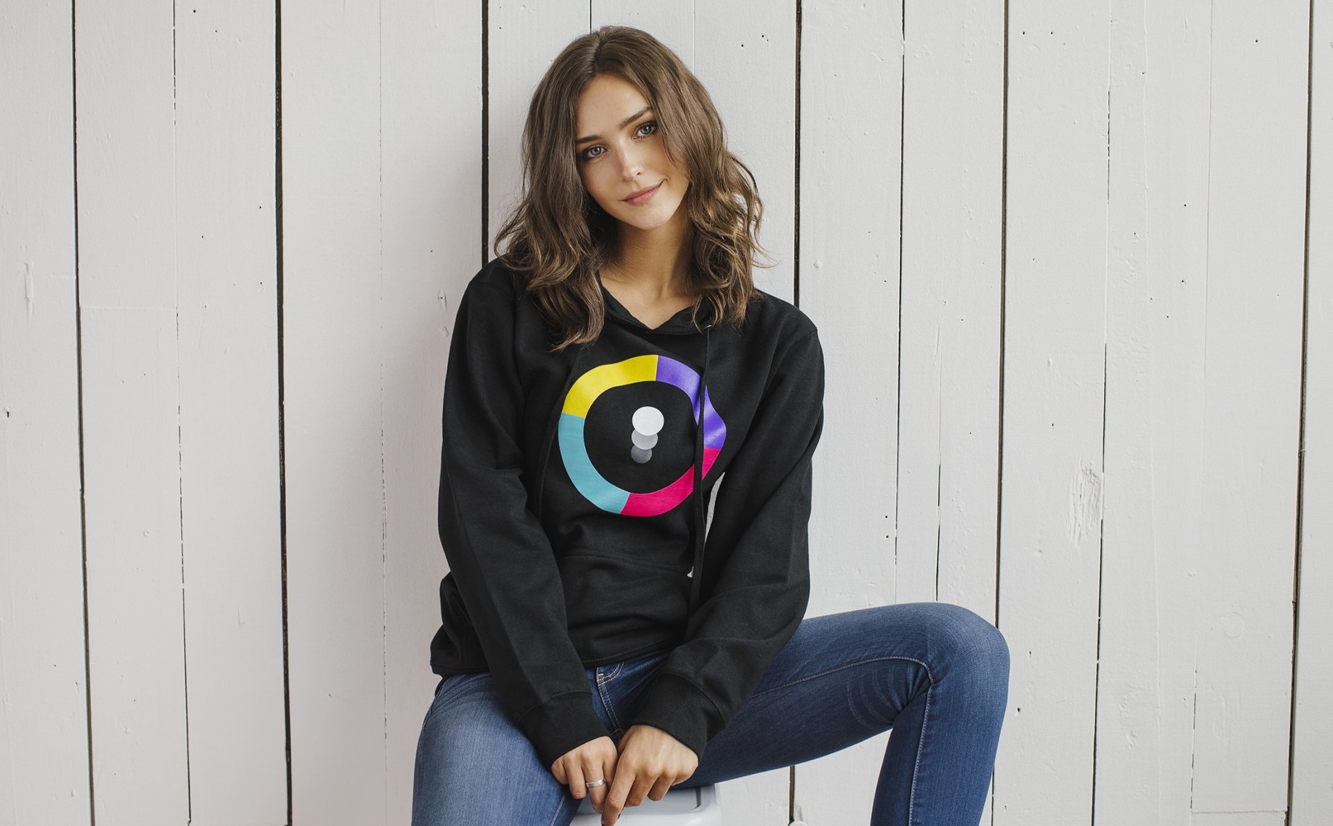 HD desktop wallpaper featuring Rachel Cook, an American brunette model, seated casually against a white wooden backdrop.