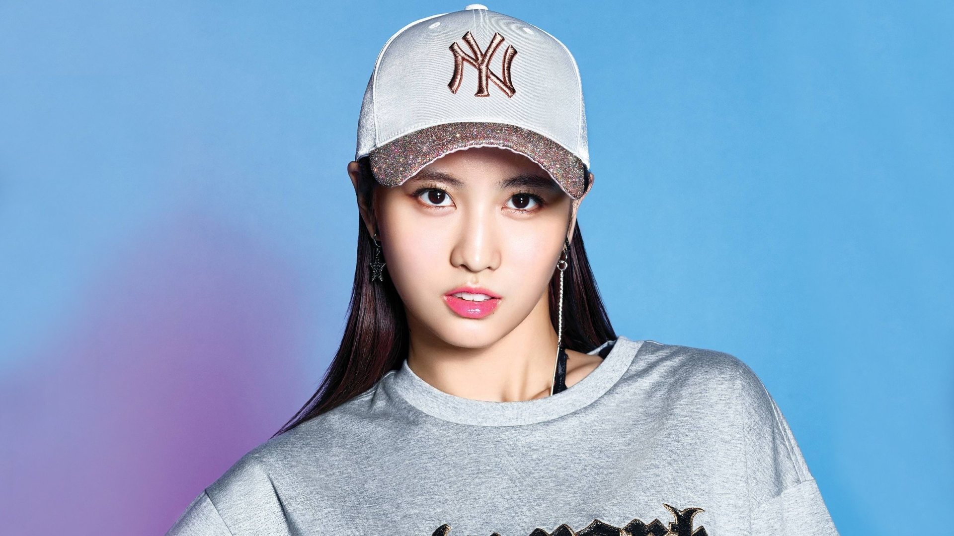 Asian woman with brown eyes wearing a New York cap and gray sweatshirt on a blue background — K-pop music–style 2K Quad HD PC desktop wallpaper.