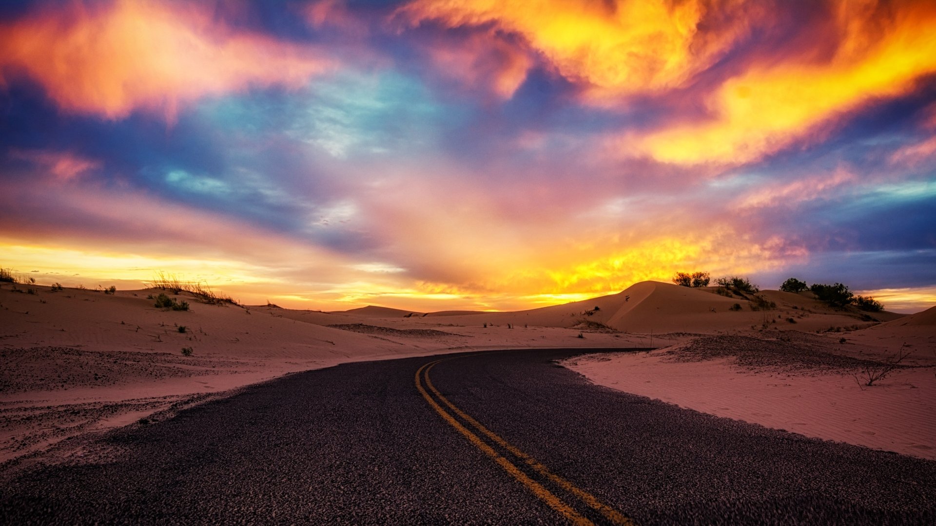 A 4K Ultra HD wallpaper featuring a man-made road winding through sand dunes under a vibrant sunset sky.