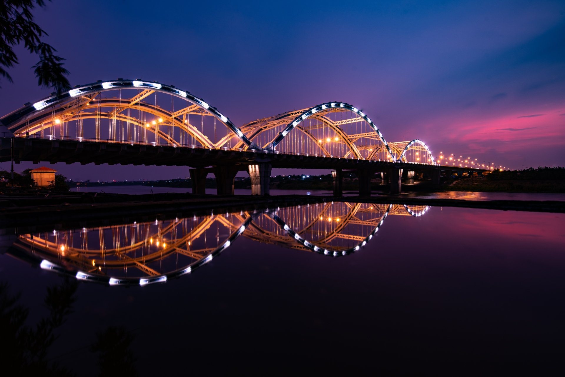 A 4K Ultra HD wallpaper showing a brightly lit arch bridge in Vietnam at night, its reflection shimmering on the calm water below under a deep twilight sky.
