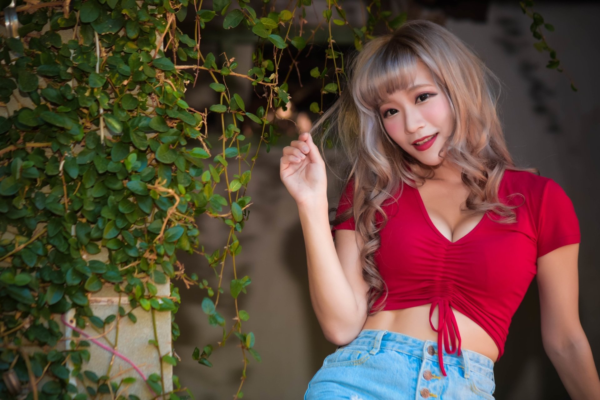 HD PC desktop wallpaper: blonde Asian model with red lipstick in a red cropped top and jeans, smiling against a lush leafy background.