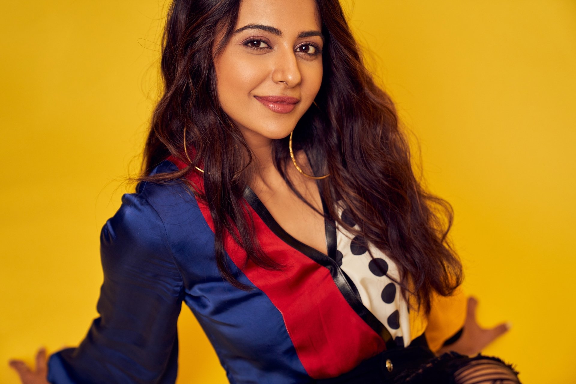 Portrait of a smiling brunette woman with long wavy hair and brown eyes against a bright yellow backdrop; 2K Quad HD PC desktop wallpaper.