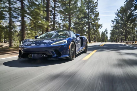 Blue McLaren GT supercar vehicle racing down a pine-lined road — 4K Ultra HD PC desktop wallpaper and background.