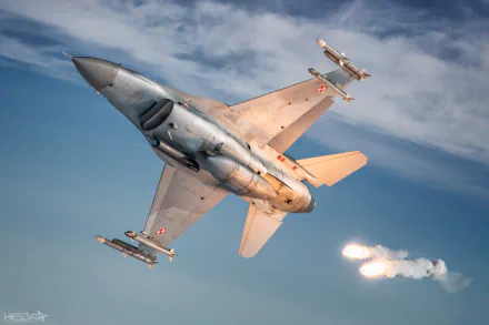 HD desktop wallpaper of a General Dynamics F-16 Fighting Falcon jet fighter warplane in flight, releasing flares against a partly cloudy sky.