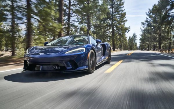 Blue McLaren GT supercar vehicle racing down a pine-lined road — 4K Ultra HD PC desktop wallpaper and background.