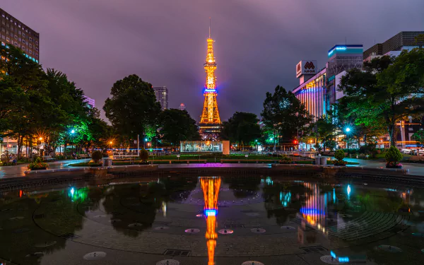 Night view of Sapporo city in Japan featuring a brightly lit tower reflected in calm water, surrounded by buildings and vibrant city lights.