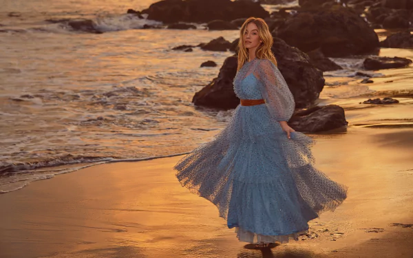 American actress Sydney Sweeney in a flowing blue dress stands on a rocky beach at sunset, captured in HD for a desktop wallpaper background.