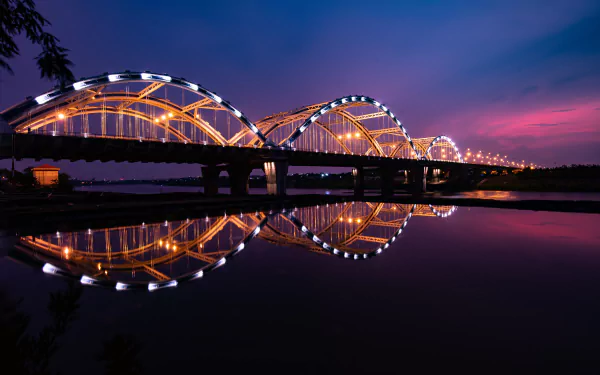 A 4K Ultra HD wallpaper showing a brightly lit arch bridge in Vietnam at night, its reflection shimmering on the calm water below under a deep twilight sky.