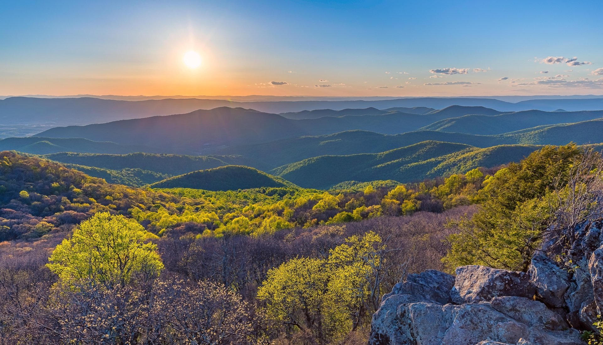 Sunset over a forested mountain landscape with bright green trees and rocky foreground, captured in vivid detail as a 4K Ultra HD desktop wallpaper.