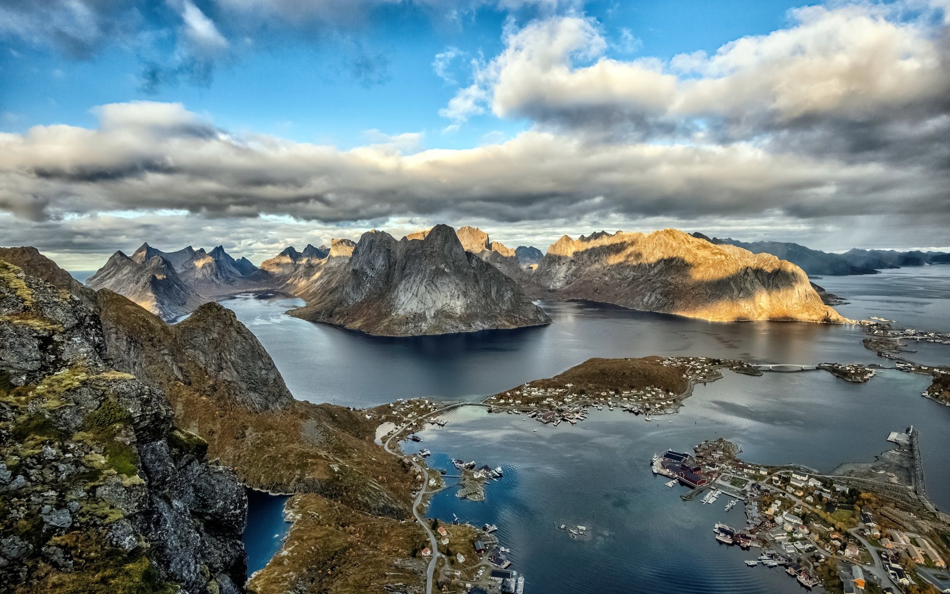 4K Ultra HD PC desktop wallpaper: Lofoten coast with jagged mountains rising from deep sea, dramatic clouds and sunlight over fjords and a small coastal village — atmospheric landscape photography.