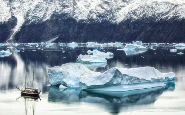HD PC desktop wallpaper and background: sailboat vehicle anchored among icebergs on glassy Greenland water, snow‑capped cliffs and floating ice reflected in the calm arctic sea.