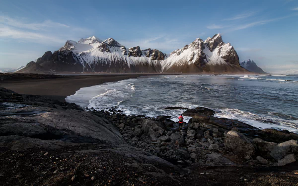 Vestrahorn Mountain rises majestically over a rocky beach in Iceland, with waves crashing and a person in red standing on the shore under a clear sky.