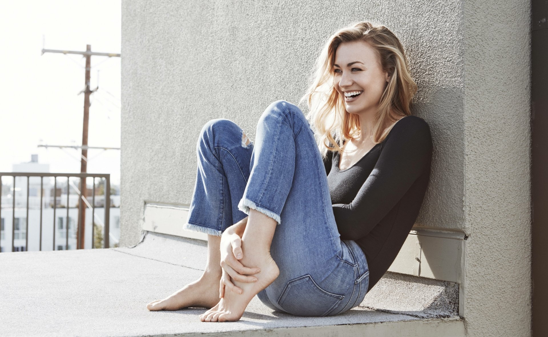 Australian actress Yvonne Strahovski smiles warmly, seated barefoot outdoors against a wall, captured in an 8K Ultra HD PC desktop wallpaper.