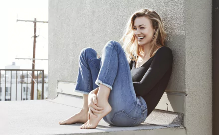 Australian actress Yvonne Strahovski smiles warmly, seated barefoot outdoors against a wall, captured in an 8K Ultra HD PC desktop wallpaper.