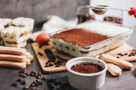 2K Quad HD PC desktop wallpaper: tiramisu still life with a glass baking dish of layered mascarpone and cocoa, ladyfingers, coffee beans and cocoa powder on a rustic board.