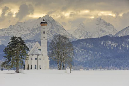 Winter scene of a religious church surrounded by trees in Schwangau, Bavaria, set against the snowy Alps mountains in Germany.