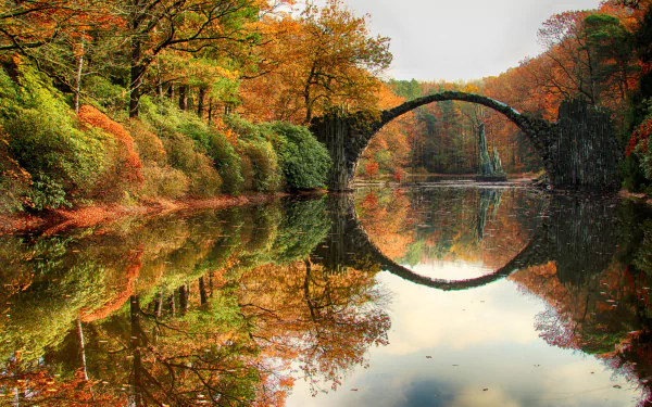 A serene autumn scene at Devil's Bridge in Germany, featuring vibrant fall foliage, a historic stone bridge, and its clear reflection in the calm river below.