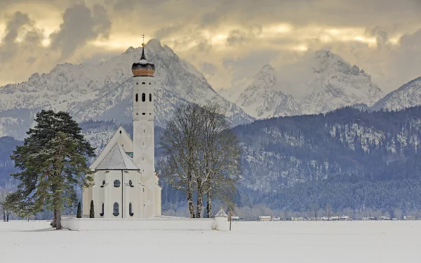 Winter scene of a religious church surrounded by trees in Schwangau, Bavaria, set against the snowy Alps mountains in Germany.