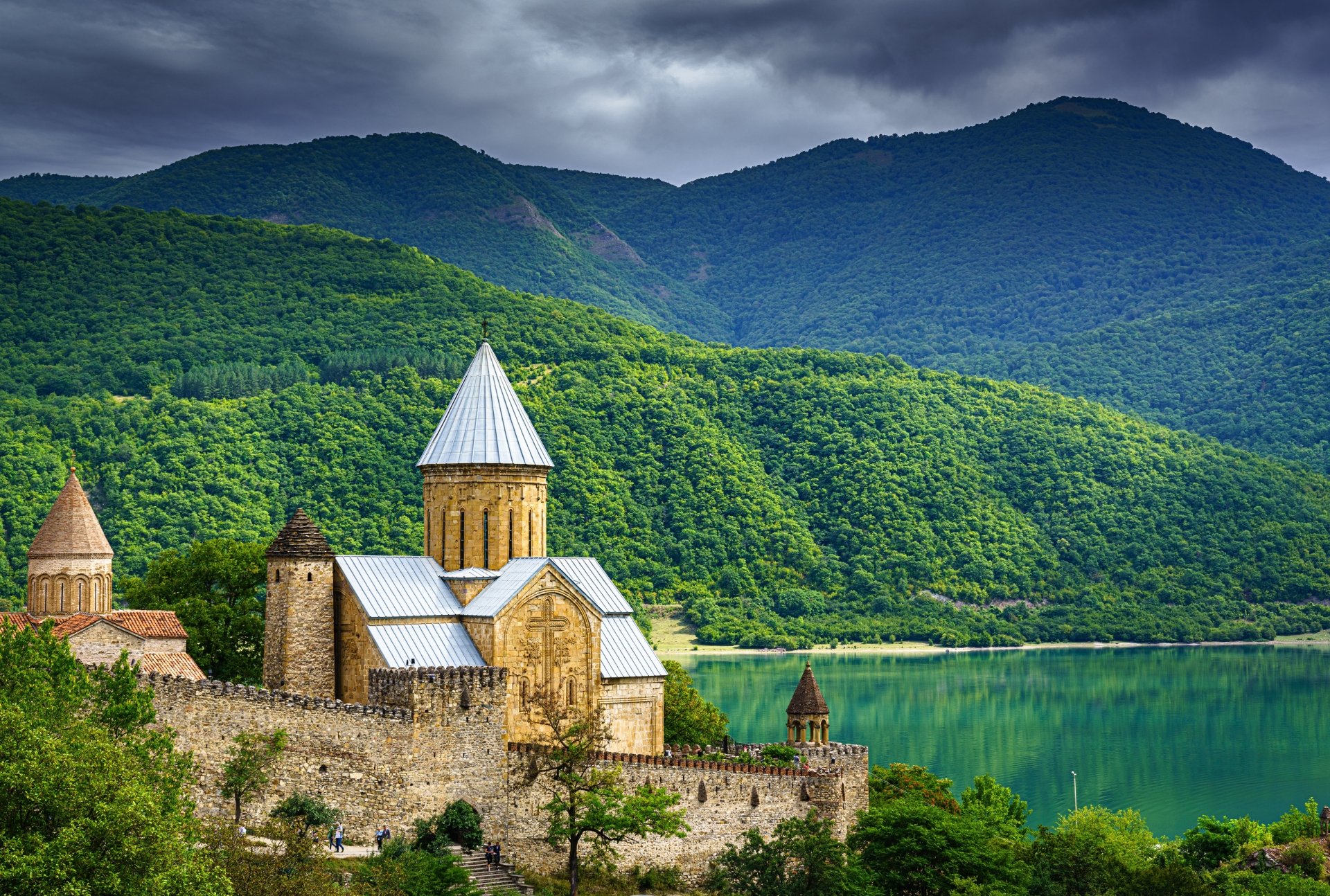 A 4K Ultra HD view of a man-made fortress and castle by a river, set against lush green mountains in Georgia under a moody sky.